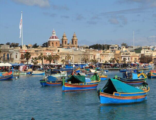 A view of a fishing village in Malta, with colourful boats in the harbour and a church in the background. Pic by Frank Vincentz under Creative Commons 3.0